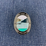 An oval enamel Walking Stick Medallion featuring Glacier Bay National Park and Preserve artwork, placed on a blue denim fabric background.