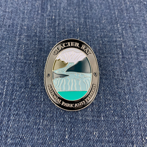 An oval enamel Walking Stick Medallion featuring Glacier Bay National Park and Preserve artwork, placed on a blue denim fabric background.