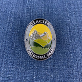 An oval enamel lapel Walking Stick Medallion featuring a mountain landscape with the text 'GLACIER NATIONAL PARK' on a blue denim background.