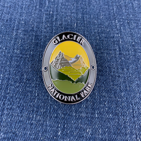 An oval enamel lapel Walking Stick Medallion featuring a mountain landscape with the text 'GLACIER NATIONAL PARK' on a blue denim background.