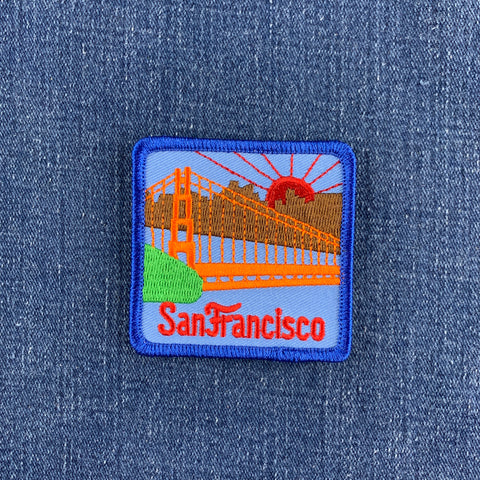 A square embroidered patch of the Golden Gate Bridge in San Francisco, featuring orange and brown bridge details, a red sun, green hill, and red 'San Francisco' text on a light blue background, stitched onto a blue denim fabric.