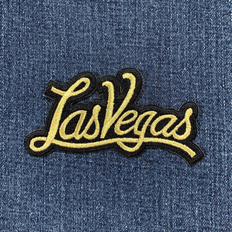 Curved embroidered patch spelling the word "Las Vegas" in glittering gold script with a black border, set against a denim background.