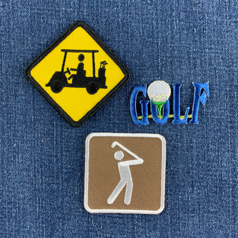 Three embroidered golf-themed patches arranged on a blue denim background: a yellow warning-sign style patch with a black golf cart silhouette, a blue 'GOLF' patch with a white golf ball and green tee detail, and a brown square patch with a white golfer silhouette.