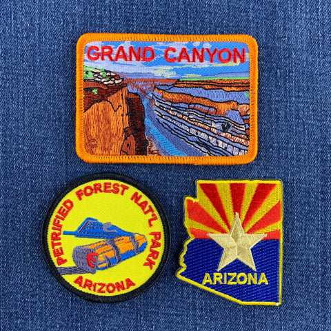 Three embroidered patches displayed on a blue denim background: a rectangular 'Grand Canyon' patch with an orange border, a round 'Petrified Forest National Park Arizona' patch with a black border, and an Arizona state-shaped patch with a yellow star and red/yellow sunburst.