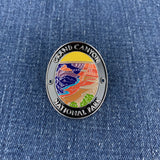 An oval enamel Walking Stick Medallion featuring the text 'GRAND CANYON NATIONAL PARK' around a colorful landscape illustration, shown on a blue denim background.