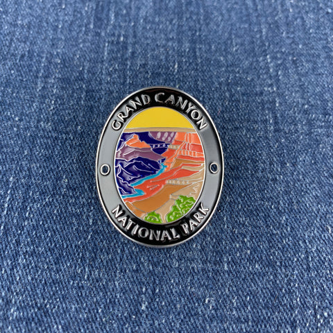 An oval enamel Walking Stick Medallion featuring the text 'GRAND CANYON NATIONAL PARK' around a colorful landscape illustration, shown on a blue denim background.