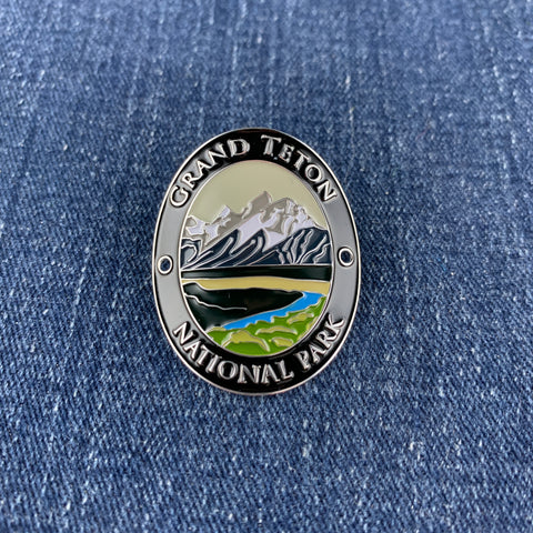 An oval Grand Teton National Park enamel Walking Stick Medallion with a silver-toned border and multicolor landscape design, placed on a blue denim background.