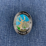 An oval metal Walking Stick Medallion featuring the Great Basin National Park emblem, resting on a blue denim fabric background. The pin has a silver-toned border with black lettering and a colorful center depicting a tree, mountains, and sky.