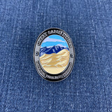 An oval enamel Walking Stick Medallion depicting the Great Sand Dunes National Park, featuring a black metal border with white lettering and a colorful landscape of blue mountains and beige sand dunes, displayed on a blue denim background.