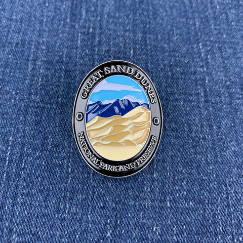 An oval enamel Walking Stick Medallion depicting the Great Sand Dunes National Park, featuring a black metal border with white lettering and a colorful landscape of blue mountains and beige sand dunes, displayed on a blue denim background.