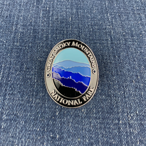 An oval enamel medallion featuring a mountain landscape design with blue, purple, and black layers, surrounded by a silver border with the text 'GREAT SMOKY MOUNTAINS NATIONAL PARK'. The pin is placed on a blue denim background.