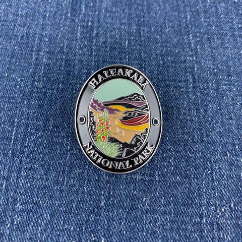 An oval enamel medallion for Haleakala National Park, featuring a detailed landscape illustration of a lake, mountains, and a bird, set against a blue denim fabric background.