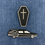 Two black embroidered patches with white outlines on a blue denim background: a coffin-shaped patch with a cross on top and a hearse car patch below.