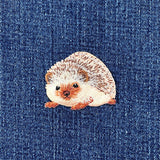 A small embroidered hedgehog patch with cream, brown, and orange tones, placed on a blue denim fabric background.
