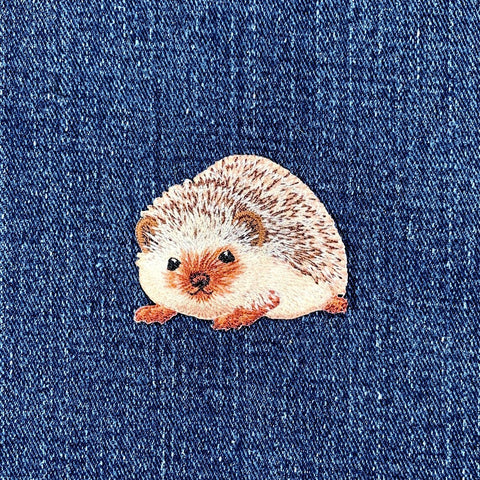 A small embroidered hedgehog patch with cream, brown, and orange tones, placed on a blue denim fabric background.