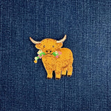 A small embroidered highland cow patch with a floral collar, placed on a textured blue denim background.