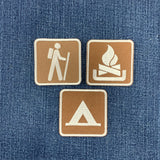 Three square embroidered patches with brown centers and white borders, featuring hiking, camping fire, and tent icons, arranged on a blue denim background.