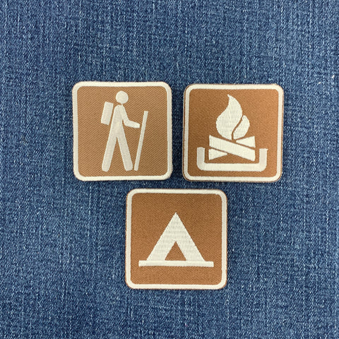 Three square embroidered patches with brown centers and white borders, featuring hiking, camping fire, and tent icons, arranged on a blue denim background.