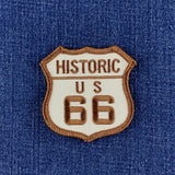 An embroidered shield-shaped patch reading 'HISTORIC US 66' in brown thread on a cream background, placed on blue denim fabric.