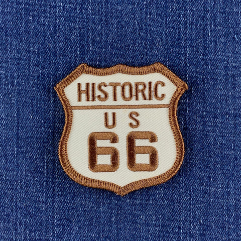 An embroidered shield-shaped patch reading 'HISTORIC US 66' in brown thread on a cream background, placed on blue denim fabric.
