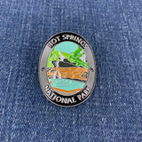 An oval enamel Walking Stick Medallion featuring a colorful landscape of Hot Springs National Park, displayed on a blue denim fabric background.