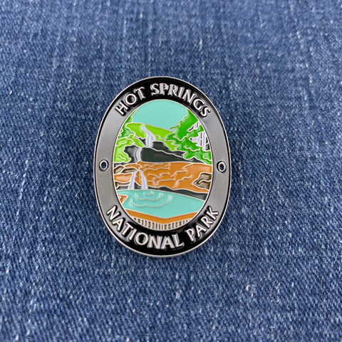 An oval enamel Walking Stick Medallion featuring a colorful landscape of Hot Springs National Park, displayed on a blue denim fabric background.