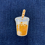 An embroidered patch shaped like a clear plastic cup filled with orange-yellow iced coffee, with a striped straw, shown on a blue denim background.