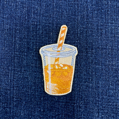 An embroidered patch shaped like a clear plastic cup filled with orange-yellow iced coffee, with a striped straw, shown on a blue denim background.