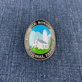 An oval enamel Walking Stick Medallion for Isle Royale National Park, featuring a lighthouse and white building on green grass under a blue sky, set against a blue denim background.