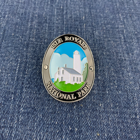 An oval enamel Walking Stick Medallion for Isle Royale National Park, featuring a lighthouse and white building on green grass under a blue sky, set against a blue denim background.