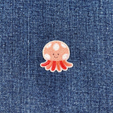 A small embroidered patch depicting a smiling jellyfish with pink tentacles and a white body with silver accents on a denim background