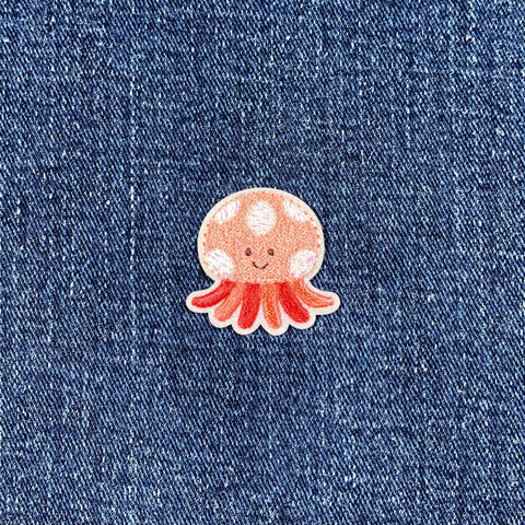 A small embroidered patch depicting a smiling jellyfish with pink tentacles and a white body with silver accents on a denim background