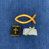 Three embroidered patches on a blue denim fabric background: a gold Ichthys fish patch, a black Holy Bible book patch with gold embroidery, and an open bible patch with a gold cross on a white page.