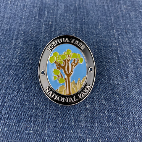 An oval enamel Walking Stick Medallion featuring the Joshua Tree National Park logo, displayed on a blue denim fabric background.