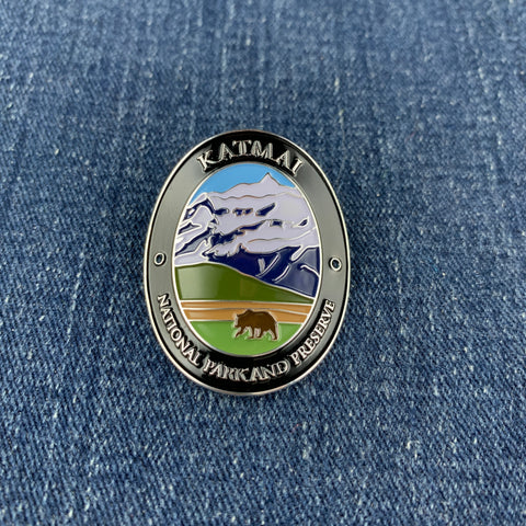 An oval metal Walking Stick Medallion featuring a scenic mountain landscape with a bear and the text 'KATMAI NATIONAL PARK AND PRESERVE', placed on a blue denim background.