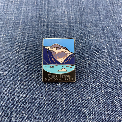 A pin badge featuring a mountain range with snow on the peaks, and the text 'Kenai Fjords National Park' on a denim background