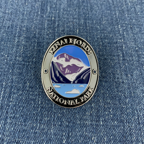 An oval enamel Walking Stick Medallion featuring Kenai Fjords National Park, displayed on a blue denim background.