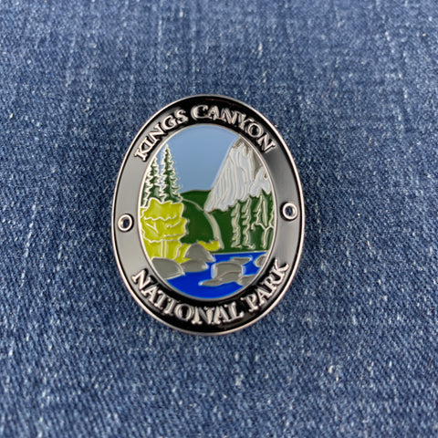 An oval metal enamel Walking Stick Medallion featuring a landscape of Kings Canyon National Park with a mountain, trees, and river, bordered by the text 'KINGS CANYON NATIONAL PARK' on a blue denim background.