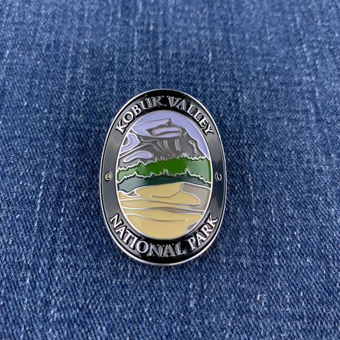 An oval metal Walking Stick Medallion featuring a landscape design of Kobuk Valley National Park, displayed on a blue denim fabric background.