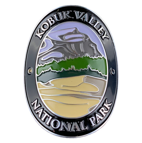 Oval Kobuk Valley National Park medallion with a black border, silver lettering, and a multicolor landscape design featuring gray mountains, green hills, and beige foreground on a white background.