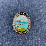 An oval enamel Walking Stick Medallion featuring a scenic landscape of Lake Clark National Park and Preserve, displayed on a blue denim fabric background.