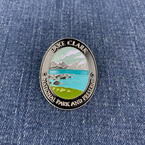An oval enamel Walking Stick Medallion featuring a scenic landscape of Lake Clark National Park and Preserve, displayed on a blue denim fabric background.