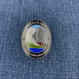 An oval Lake Clark National Park and Preserve challenge Walking Stick Medallion with a silver-toned border and multicolor landscape design, placed on a blue denim fabric background.