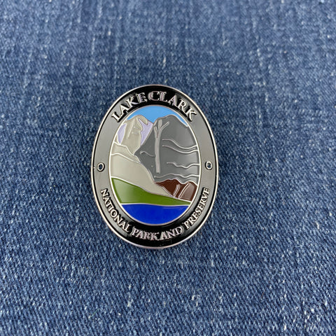 An oval Lake Clark National Park and Preserve challenge Walking Stick Medallion with a silver-toned border and multicolor landscape design, placed on a blue denim fabric background.
