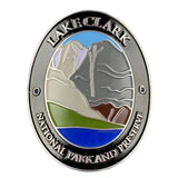 An oval metal Walking Stick Medallion for Lake Clark National Park and Preserve, featuring a colorful landscape design with mountains, water, and grass, surrounded by a silver border with raised text.