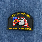 An embroidered military-style patch featuring a bald eagle over an American flag, with the text 'LAND OF THE FREE BECAUSE OF THE BRAVE,' shown on a blue denim background.
