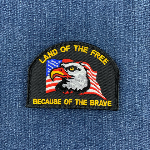 An embroidered military-style patch featuring a bald eagle over an American flag, with the text 'LAND OF THE FREE BECAUSE OF THE BRAVE,' shown on a blue denim background.