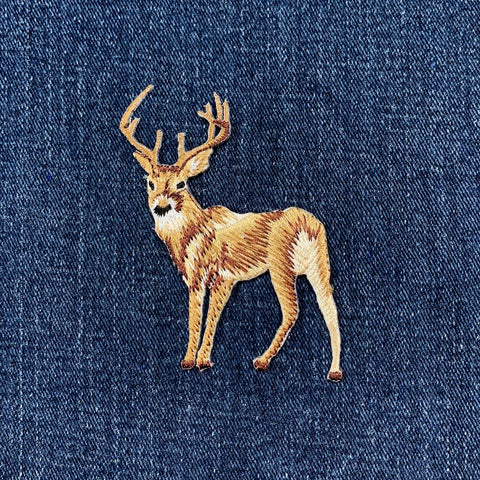 A close-up of a blue denim fabric featuring an embroidered deer patch in tan, brown, and cream tones.