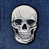 A white and black embroidered skull patch with a black border, shown on a blue denim background.