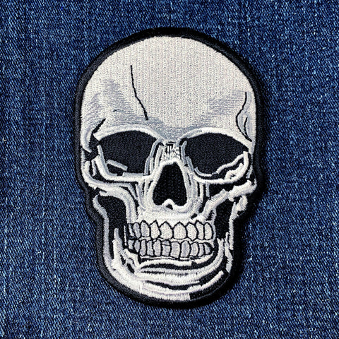A white and black embroidered skull patch with a black border, shown on a blue denim background.